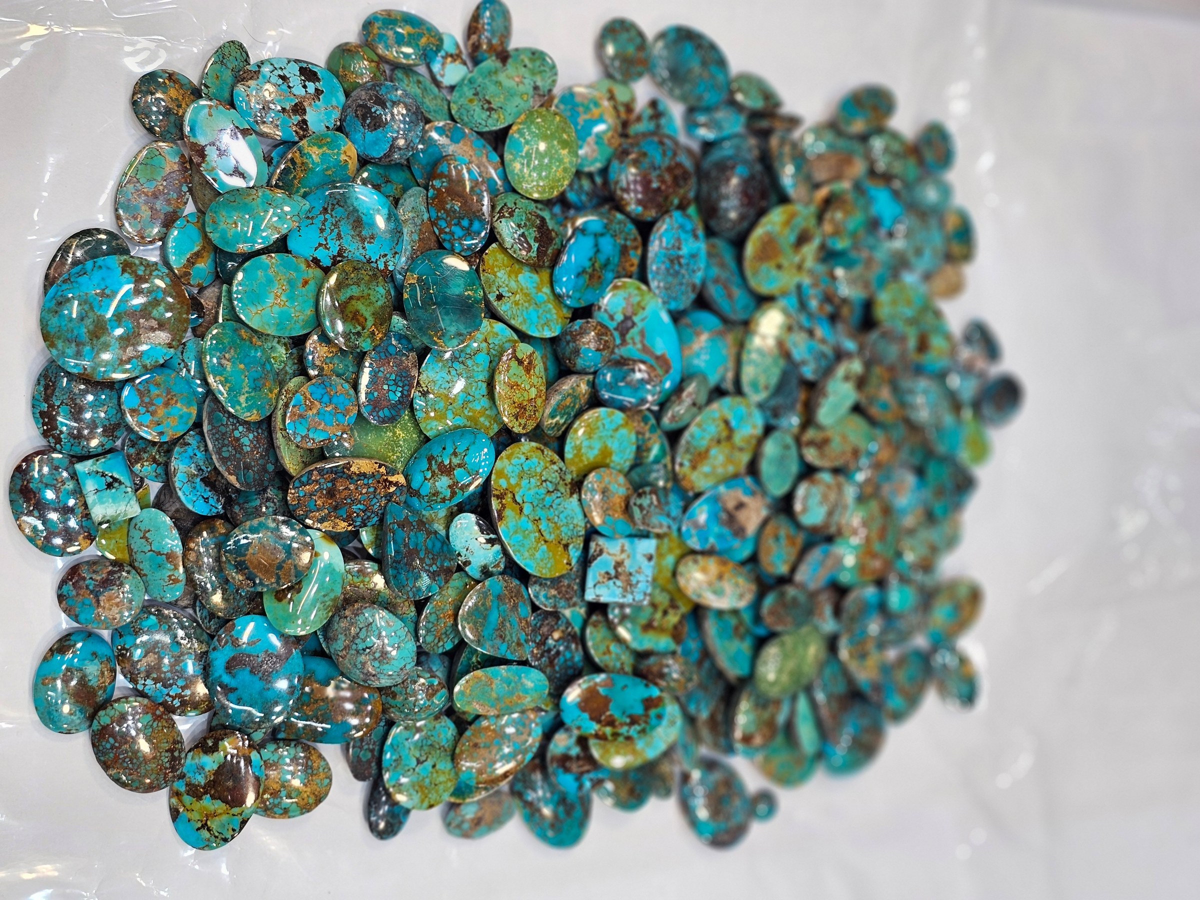 A pile of loose polished turquoise cabochons in green, brown and blue tones on a white sheet.