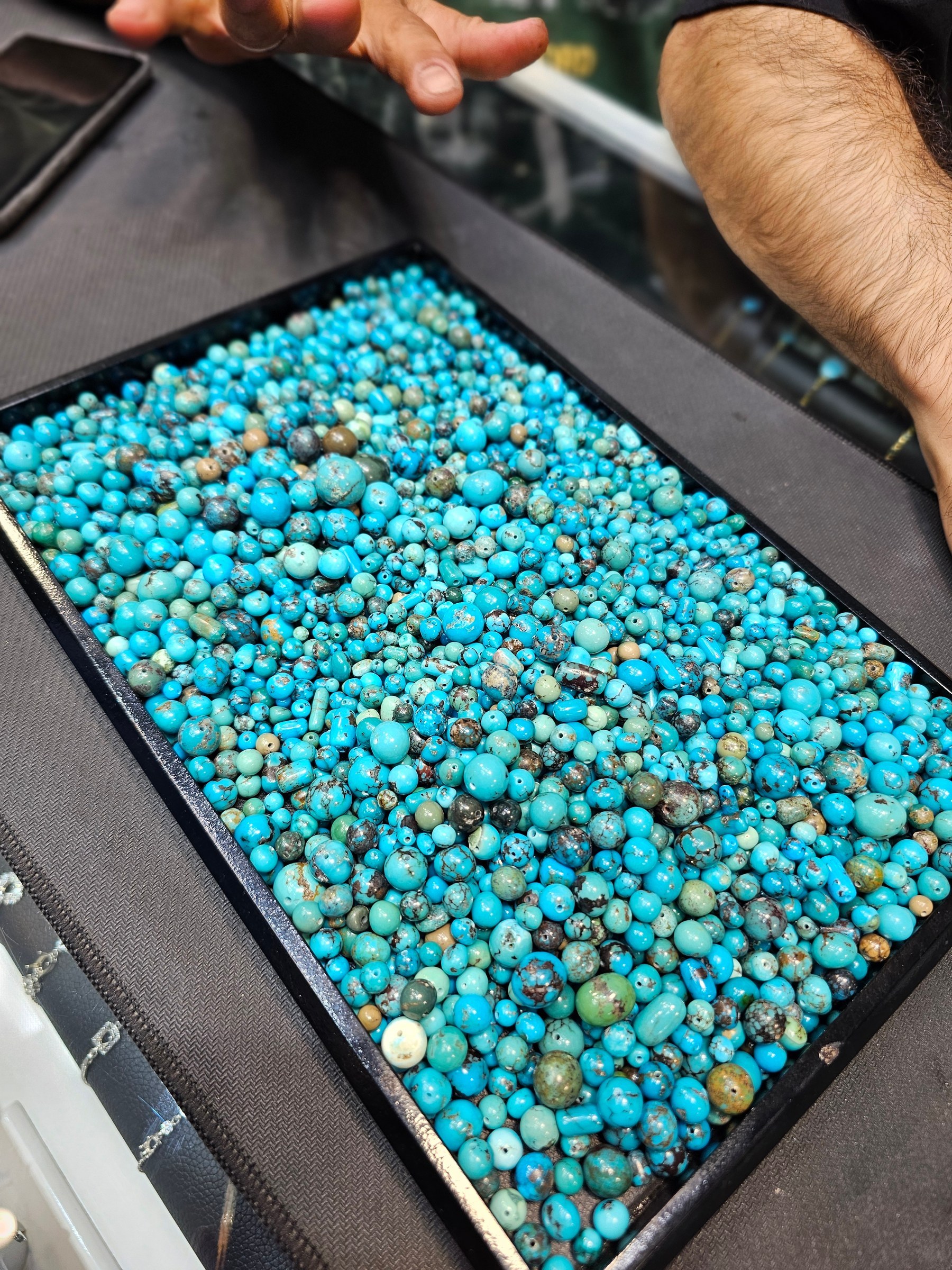 A black tray filled with small round drilled turquoise beads in bright blue with green matrix.
