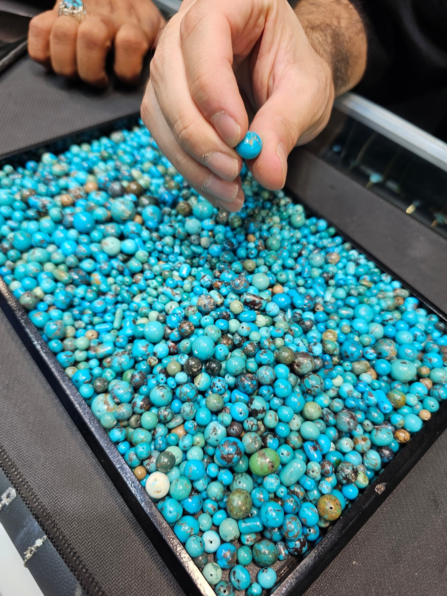 A hand picks up a single small round drilled turquoise bead from a tray of blue beads.
