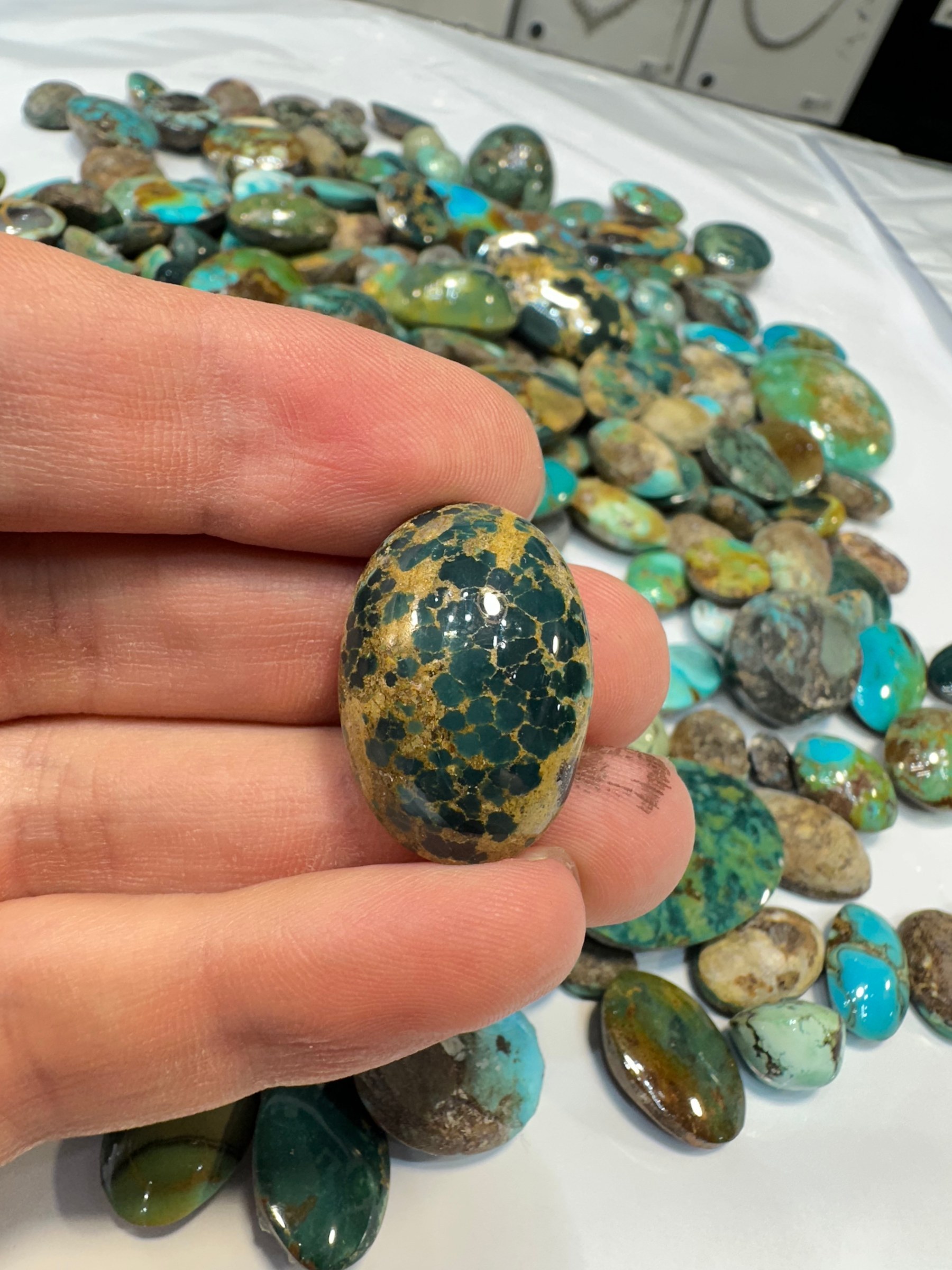 A hand holds a polished turquoise cabochon in green-brown with dark matrix beside a parcel.