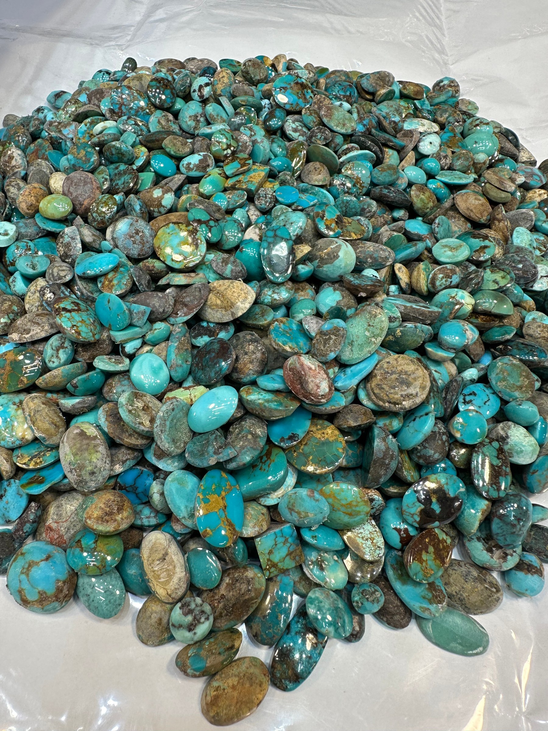 A wide spread of loose polished turquoise cabochons in green and brown tones on a white sheet.